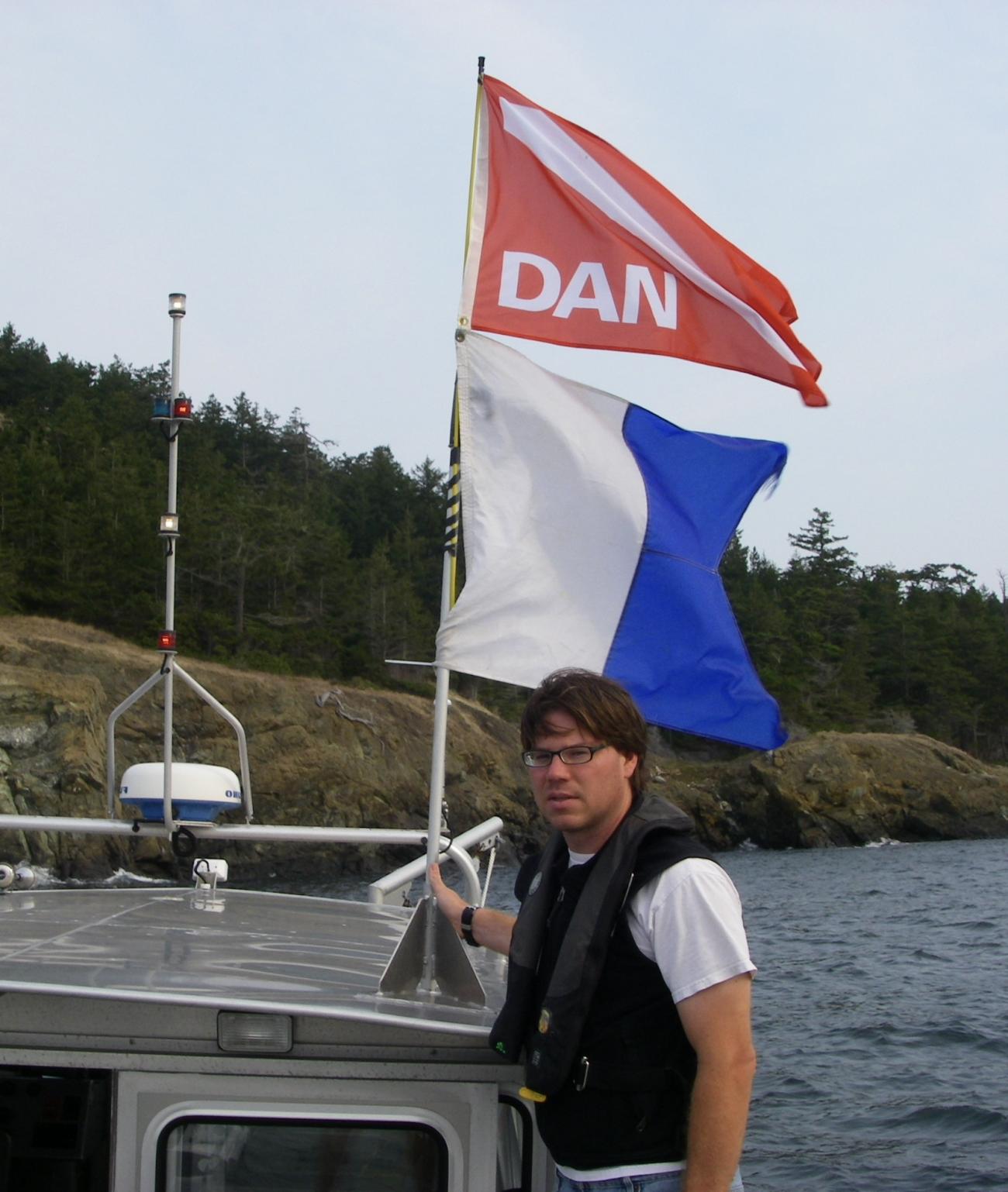 A man standing on a boat holds two flags, one red/white and one white/blue.